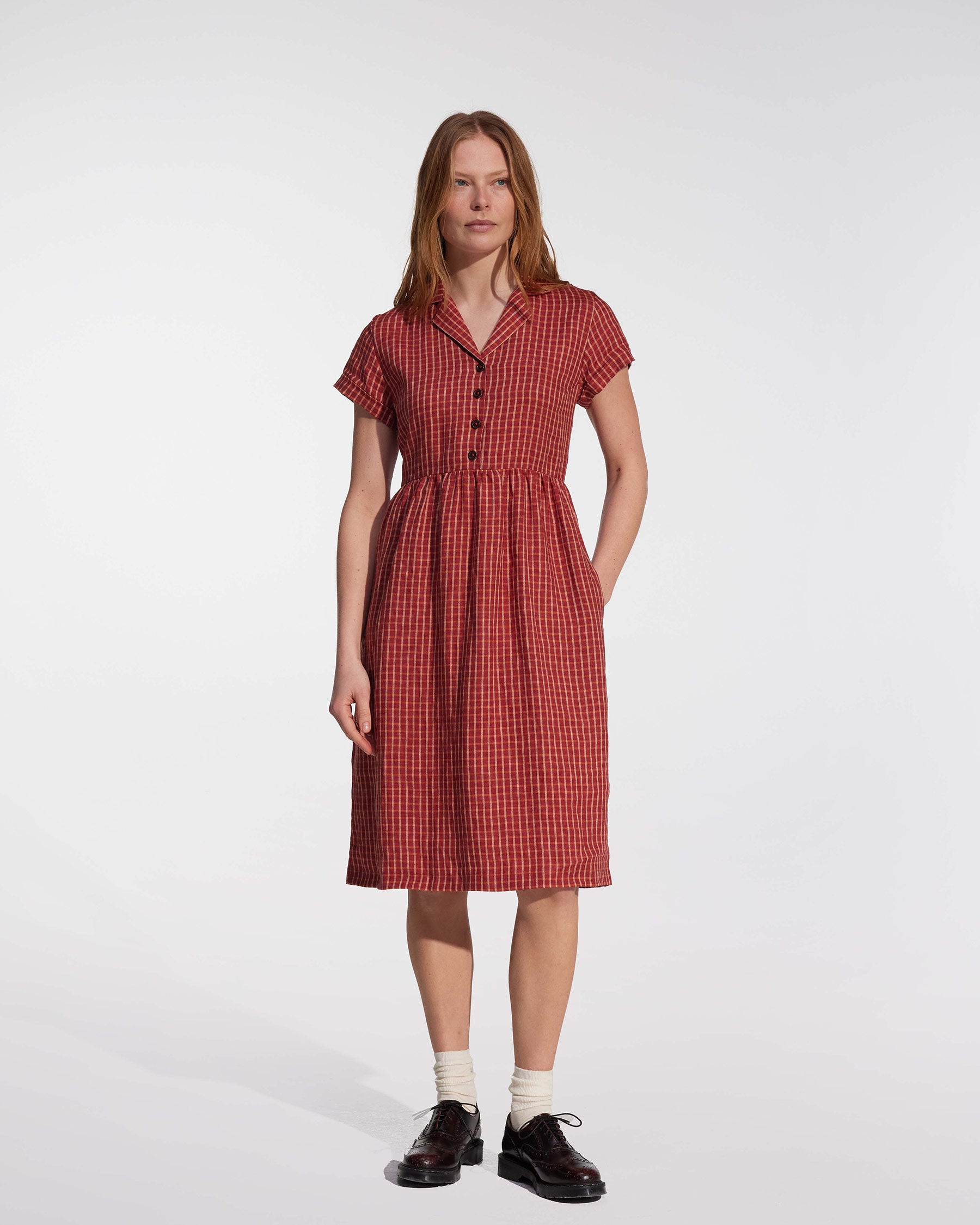 A woman stands against a plain white background wearing the No.30 Collared Linen Dress in Pomodoro Plaid by Pyne and Smith Clothiers. She has straight hair, white socks, black shoes, and one hand in her pocket.