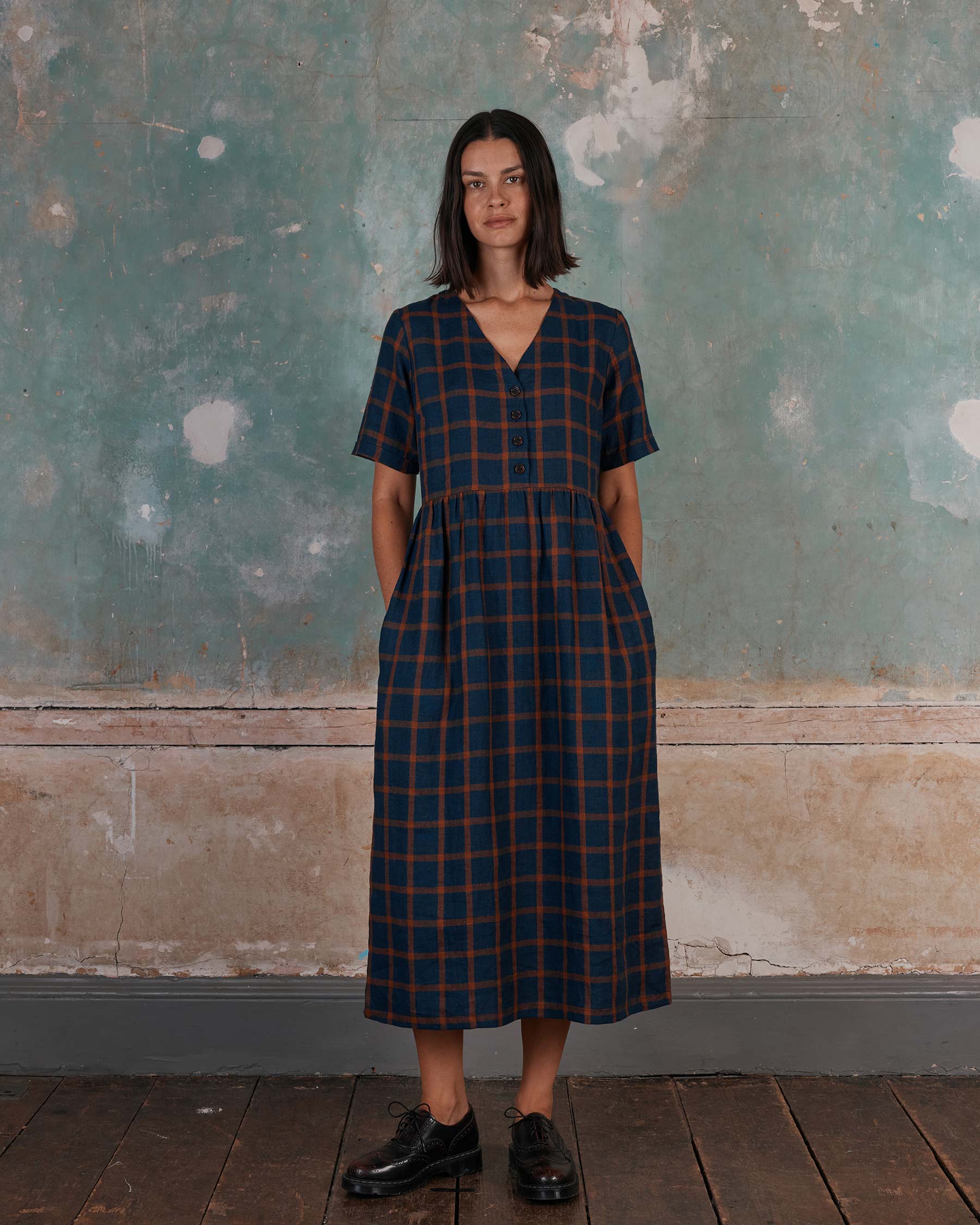 A woman stands indoors by a textured wall, wearing the Pyne and Smith Clothiers No.31 V Neck, Full Length Linen Dress in Montague Windowpane—navy blue with red plaid accents. She pairs it with black shoes and rests her hands casually in her pockets.