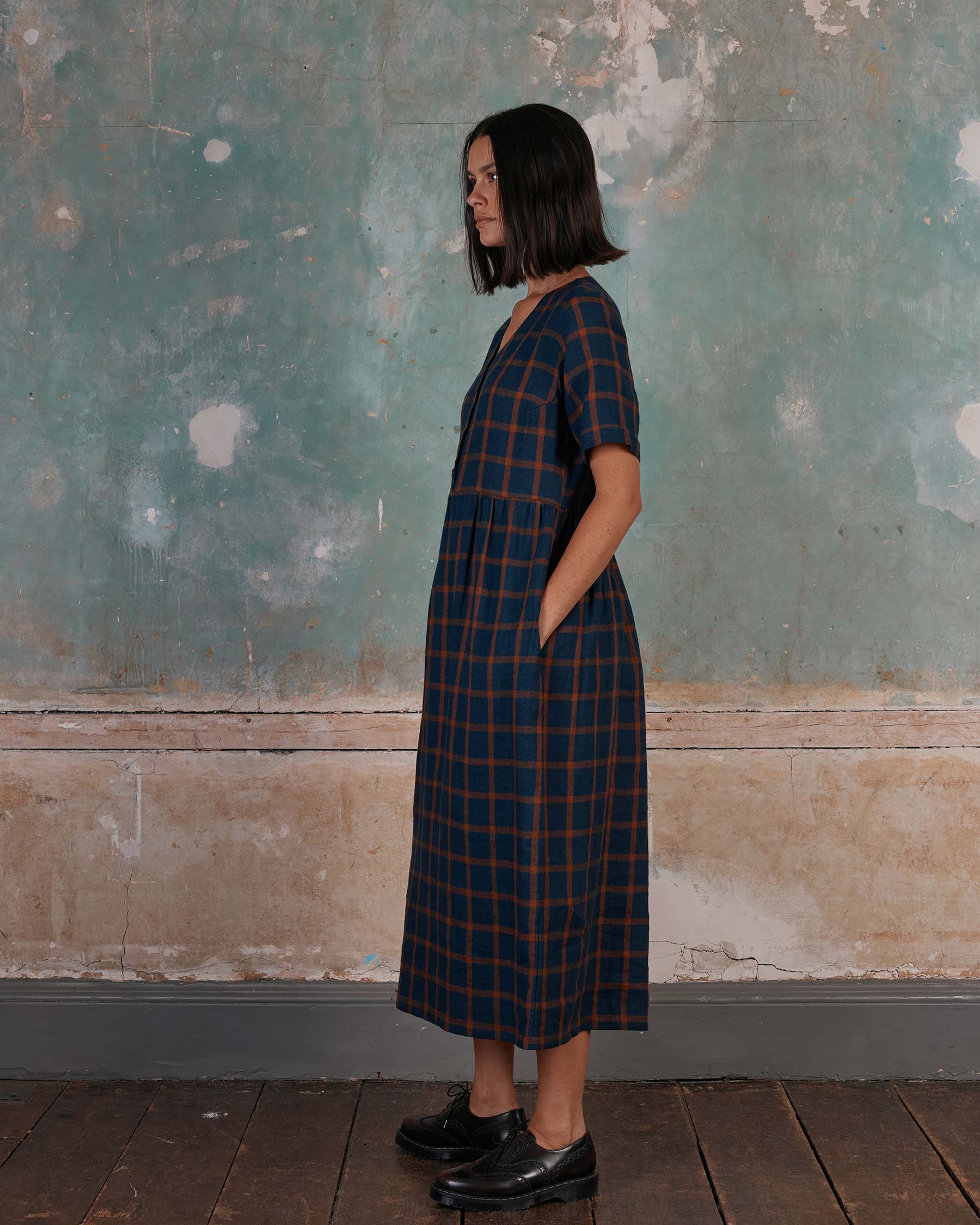 A woman with a bob stands in profile against a weathered blue-green wall, wearing the No.31 V Neck, Full Length Linen Dress in Montague Windowpane by Pyne and Smith Clothiers—an effortless California-made look with pockets and black shoes.