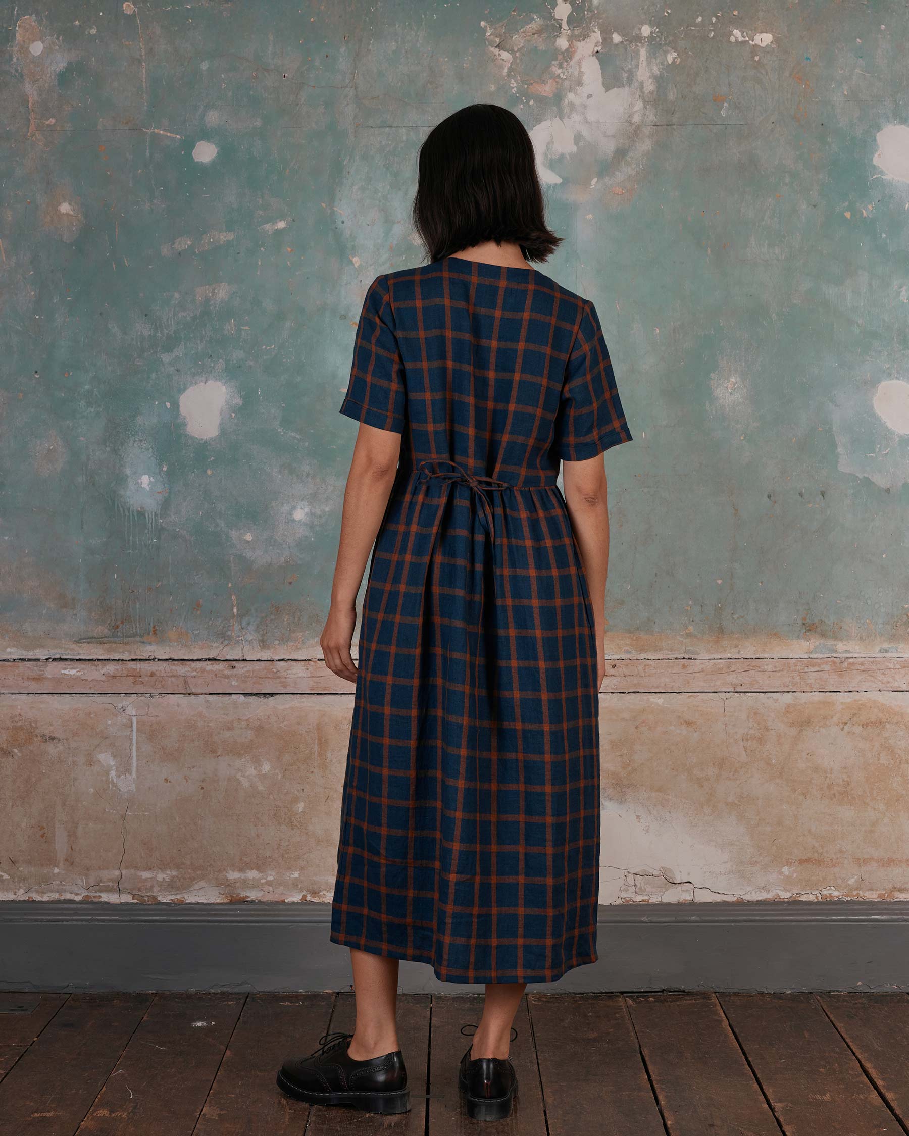 A person with short dark hair stands on a wooden floor, facing away, wearing the Pyne and Smith Clothiers No.31 V Neck, Full Length Linen Dress in Montague Windowpane and black shoes. The background is a weathered, textured wall.