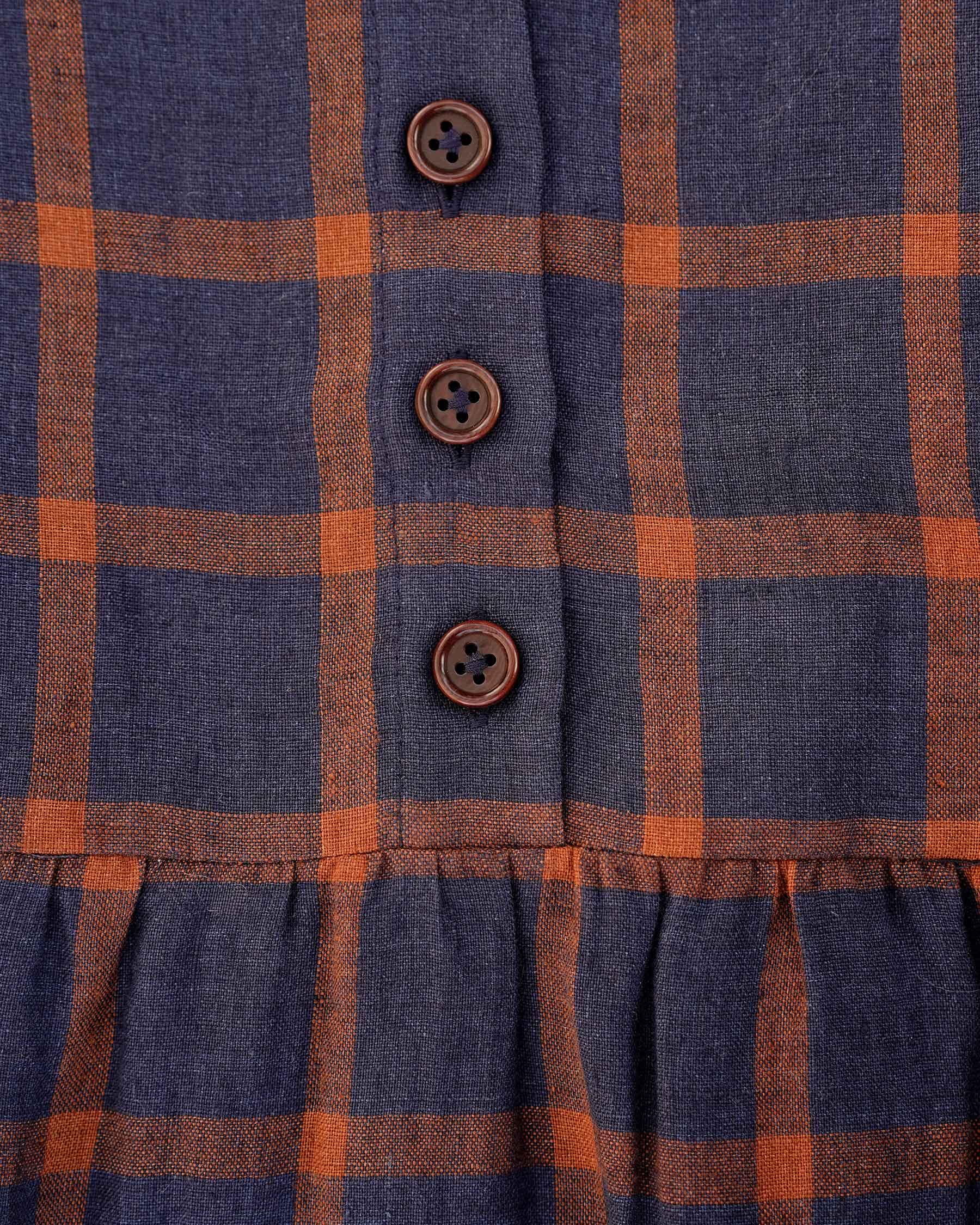 Close-up of the Pyne and Smith Clothiers No.31 V Neck, Full Length Linen Dress in Montague Windowpane, featuring an orange and dark blue plaid pattern, three brown buttons down the front, and gathered stitching at the hem.