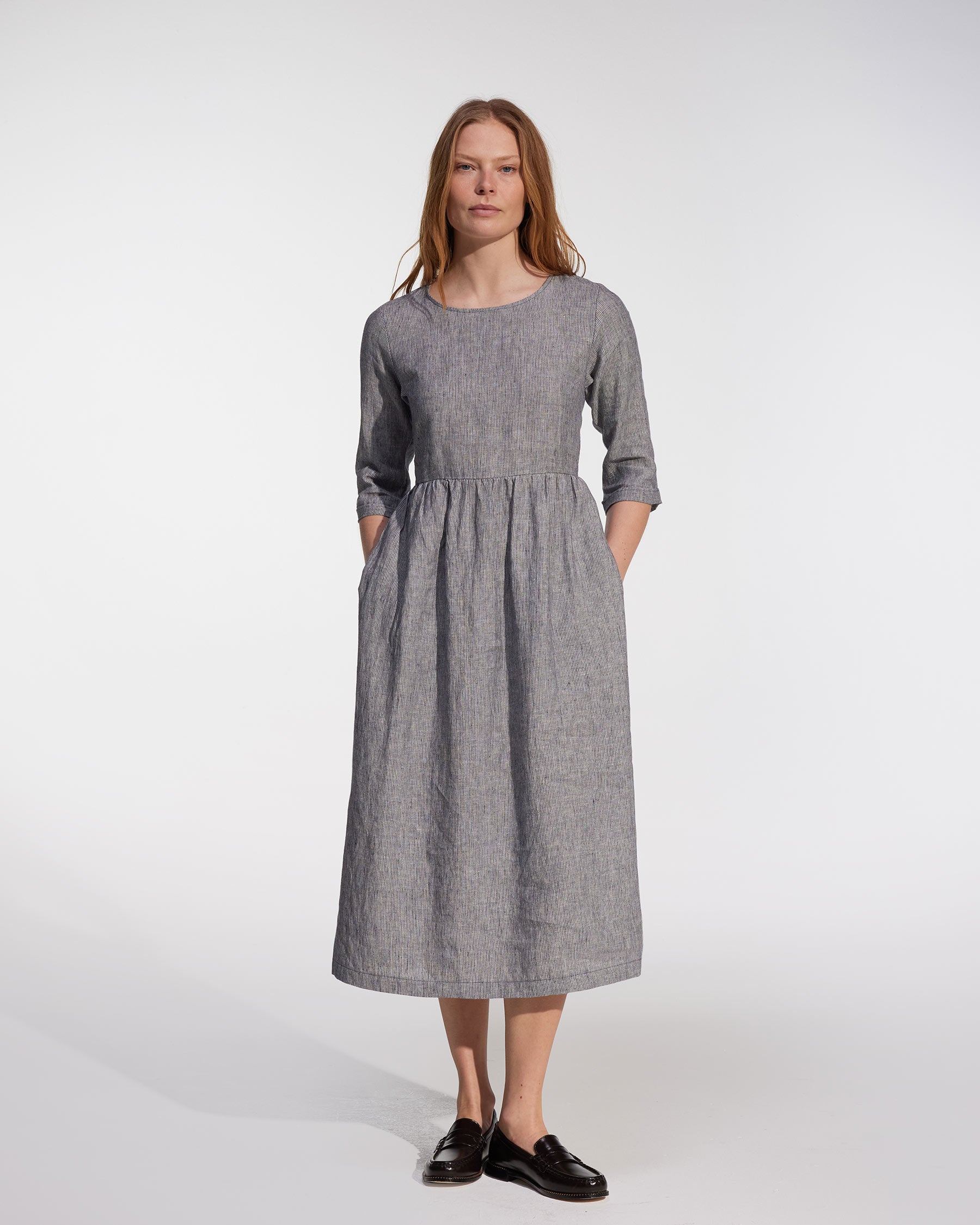 A woman with long, straight hair and a neutral expression wears the Pyne and Smith Clothiers No.41 Full Length Linen Day Dress in Cooke's Stripe, featuring long sleeves, pockets, an adjustable waist tie, and black loafers. Made in California.