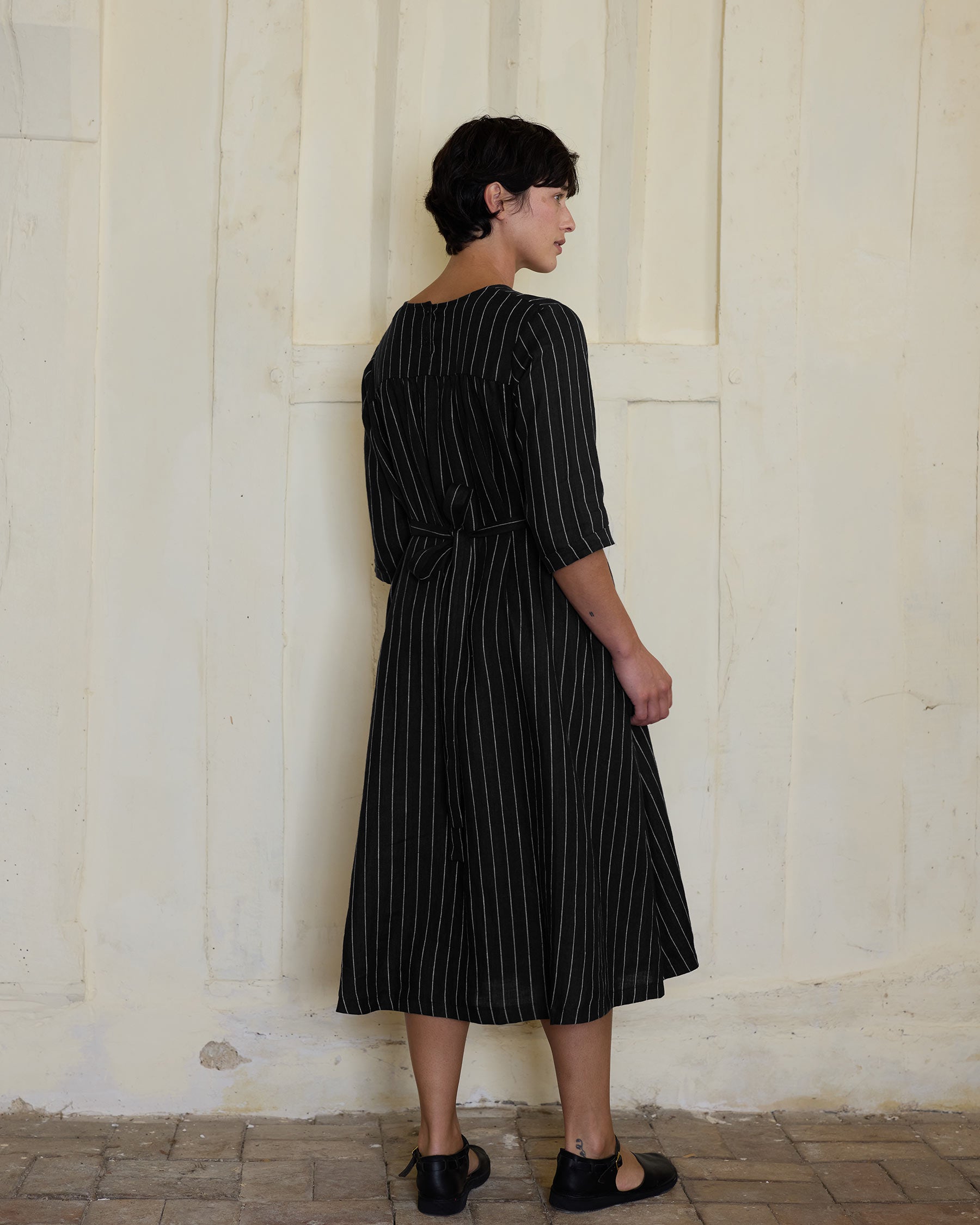 A person with short dark hair stands sideways, showcasing sustainable fashion in Pyne and Smith Clothiers No.35 Maxi Length Linen Dress in Le Marais Stripe against a light-textured wall.