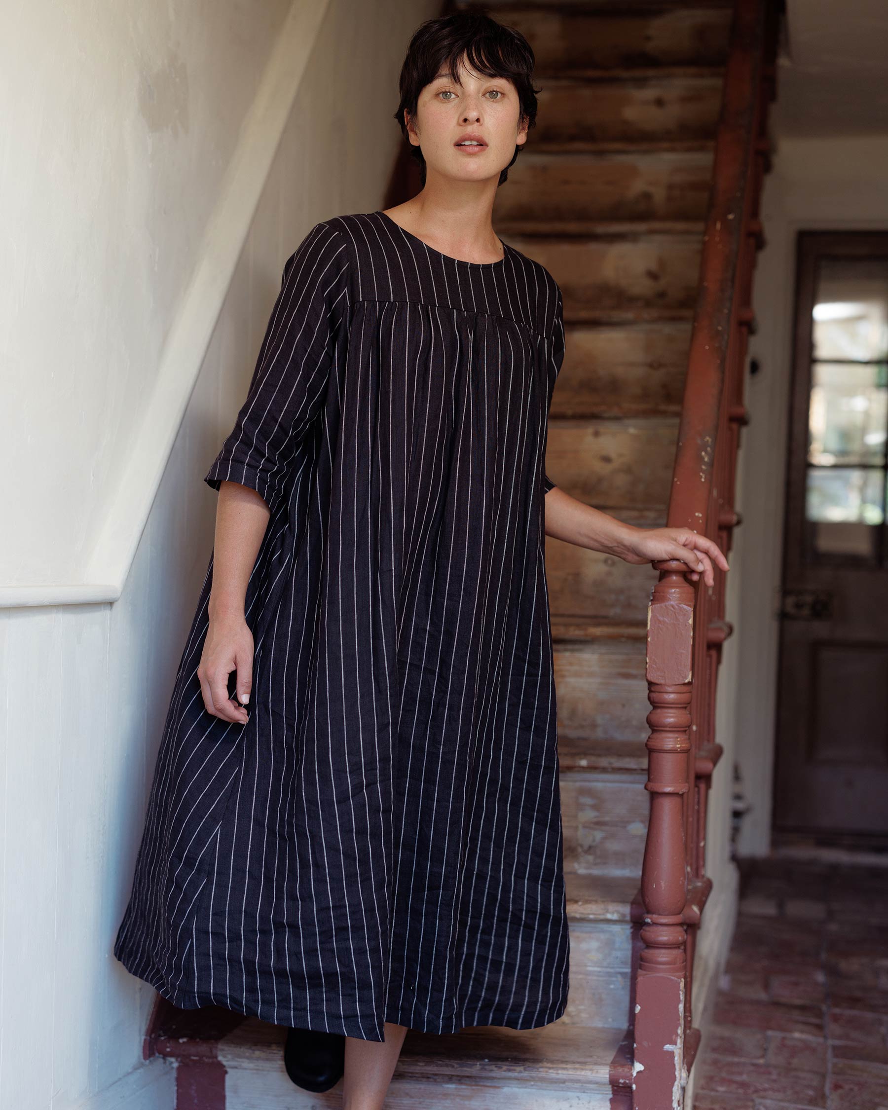 A woman with short dark hair stands indoors by a wooden staircase, wearing the Pyne and Smith Clothiers No.35 Maxi Length Linen Dress in Le Marais Stripe—an elegant, sustainable choice with timeless black and white vertical stripes.