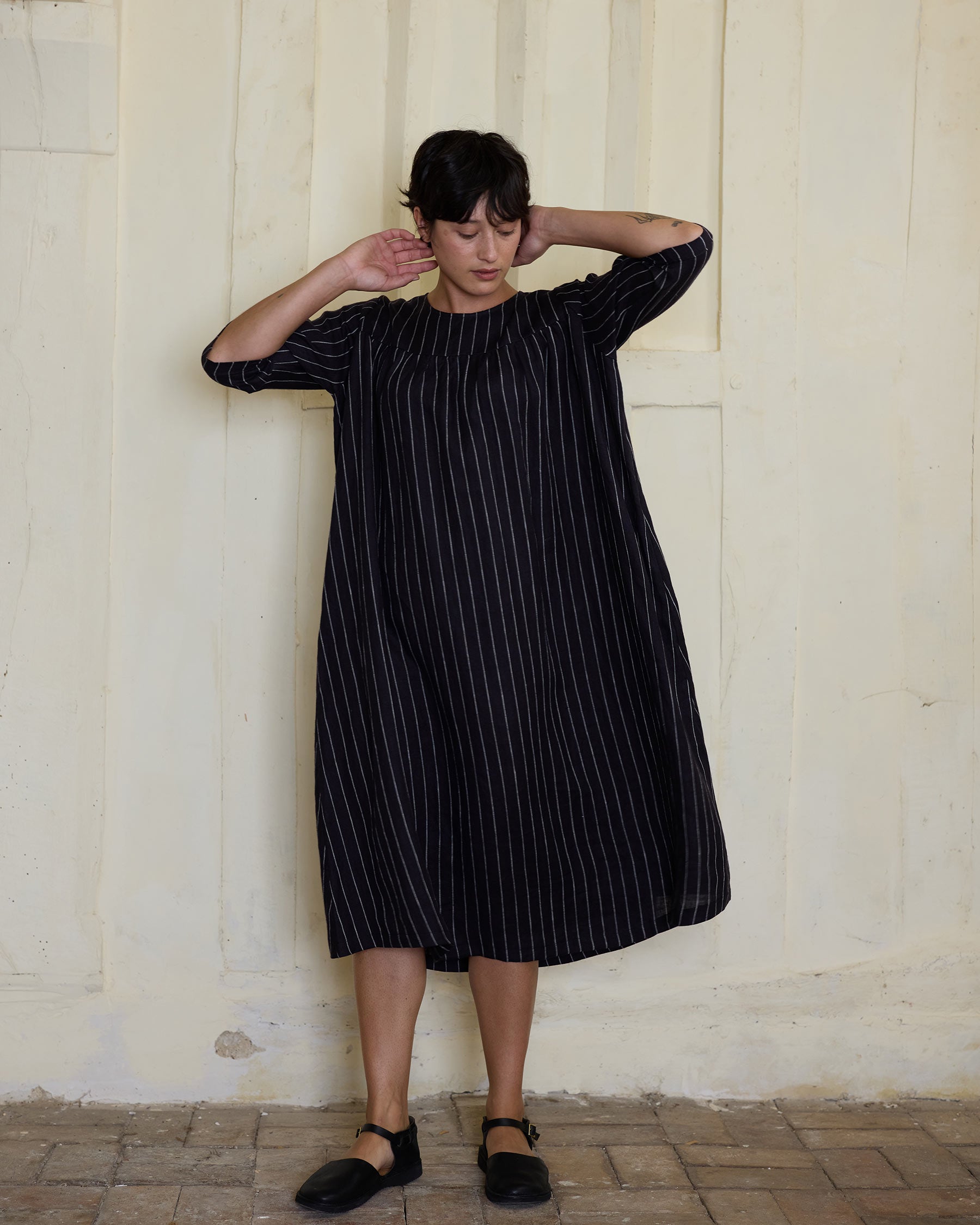 A person stands indoors by a textured wall, wearing the No.35 Maxi Length Linen Dress in Le Marais Stripe by Pyne and Smith Clothiers, paired with black shoes—an effortlessly stylish and sustainable look.