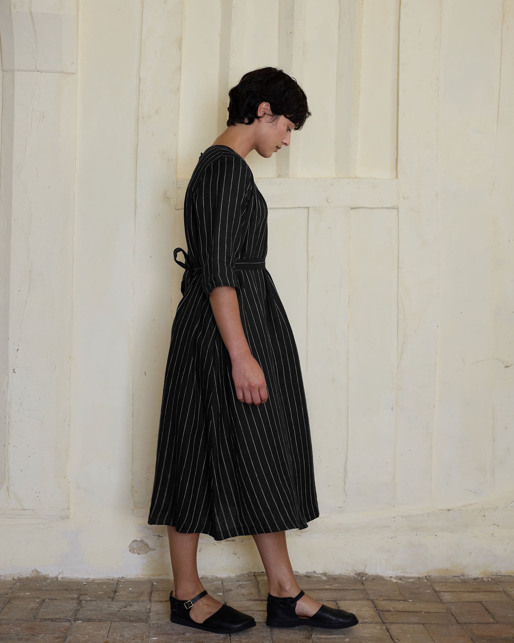A person with short dark hair stands in profile against a light, textured wall and tiled floor, wearing Pyne and Smith Clothiers No.35 Maxi Length Linen Dress in Le Marais Stripe with black shoes.