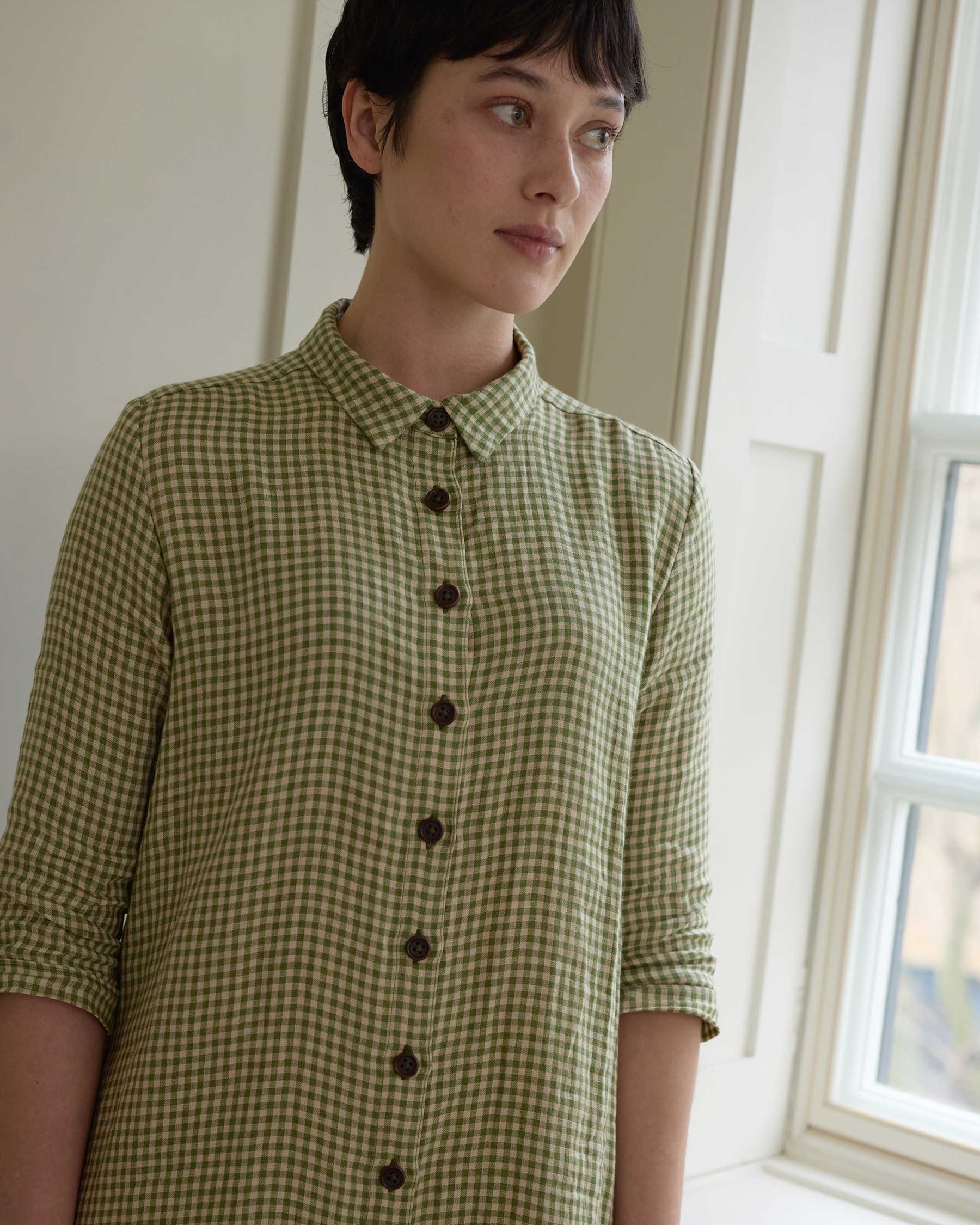 A person with short dark hair stands by a window, wearing Pyne and Smith Clothiers No.39 The Linen Shirtdress in Wimbledon—a green and white checkered linen dress with 3/4 sleeves—looking thoughtfully to the side.