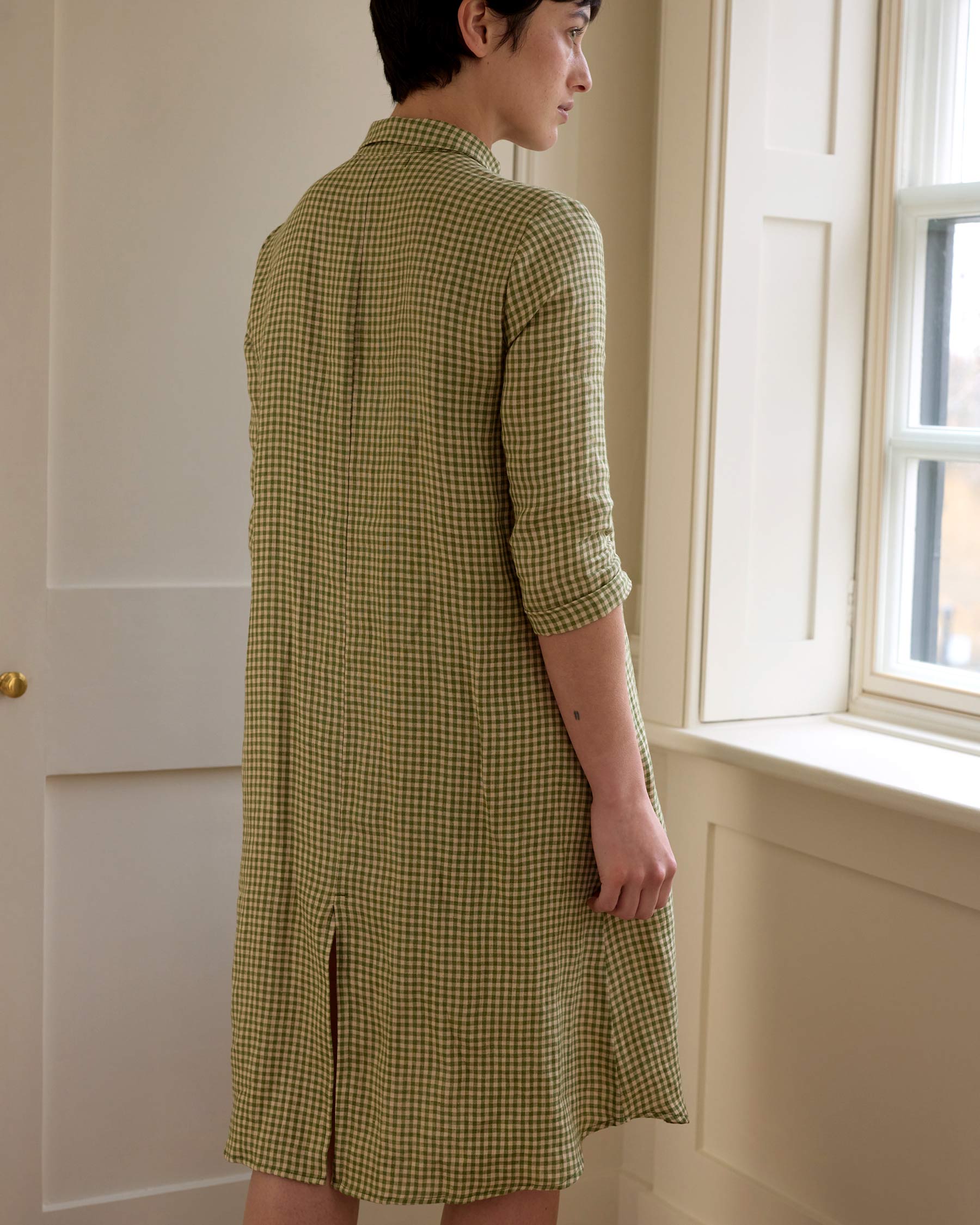 A person with short dark hair stands by a bright window, shown from behind, wearing Pyne and Smith Clothiers No.39 The Linen Shirtdress in Wimbledon—featuring green and white gingham, 3/4 sleeves, collar, back slit, and European linen.
