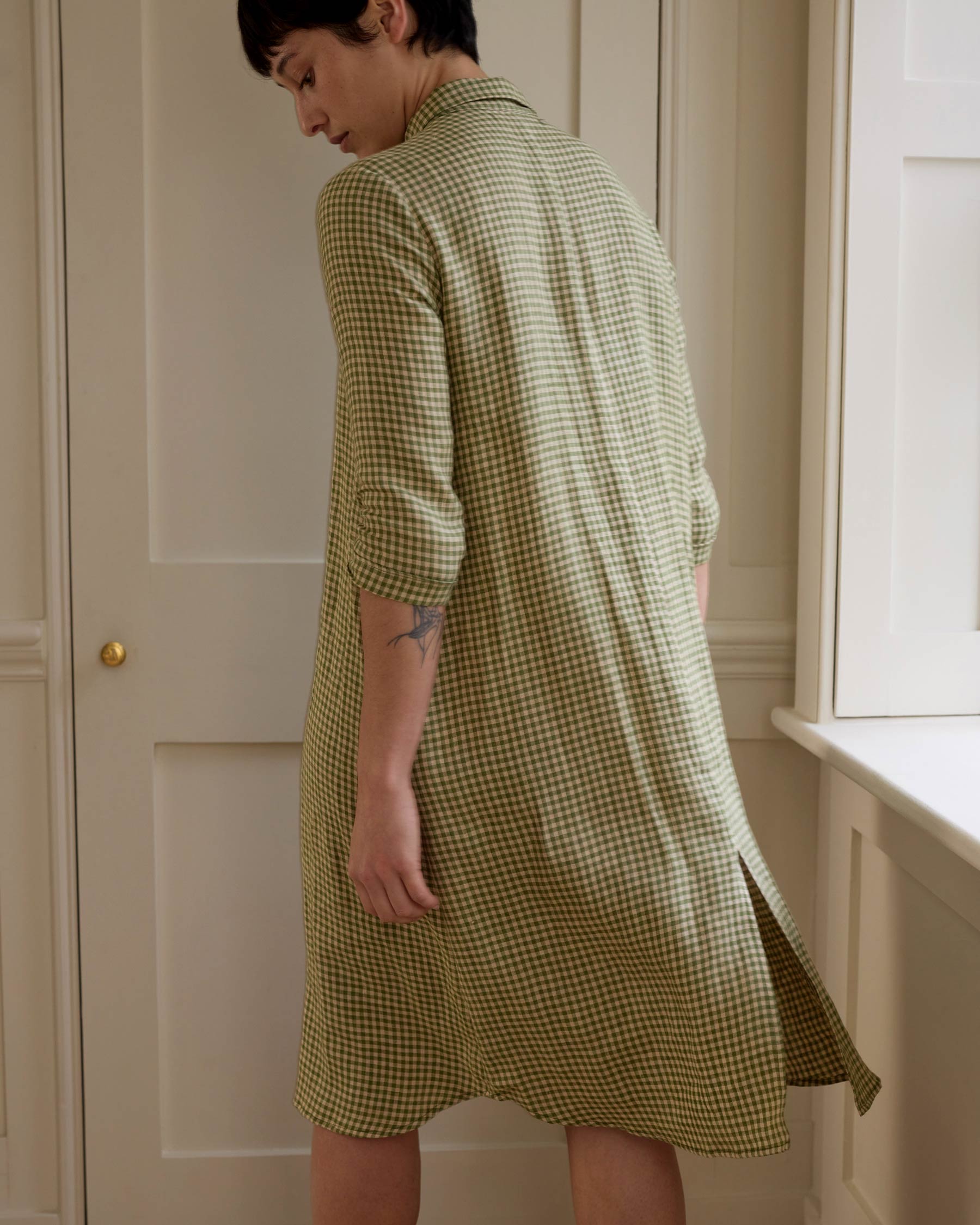 A person with short dark hair stands indoors, turned away from the camera to reveal a small left arm tattoo, wearing Pyne and Smith Clothiers No.39 The Linen Shirtdress in Wimbledon—a light green and white checkered dress with 3/4 sleeves.