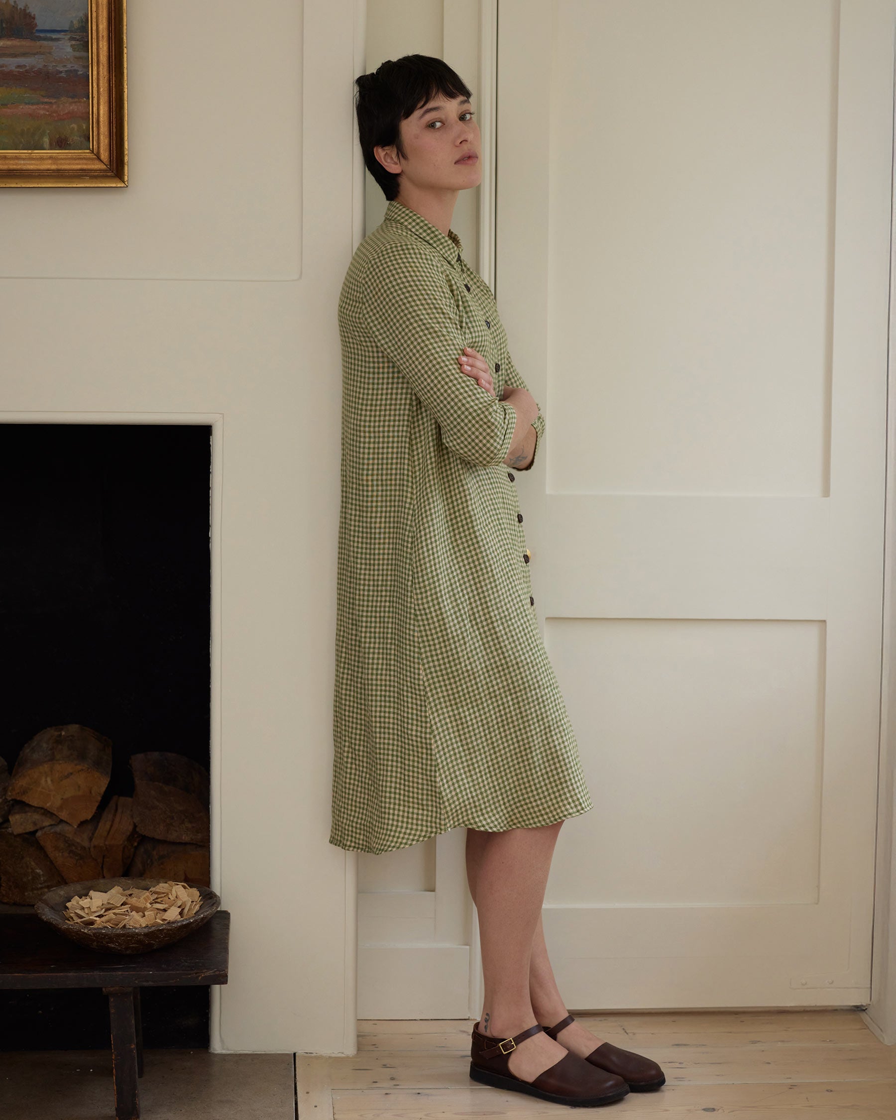 A person with short dark hair stands indoors, leaning against a cream wall with arms crossed, wearing the No.39 The Linen Shirtdress in Wimbledon by Pyne and Smith Clothiers. A fireplace with stacked wood and a painting are nearby.