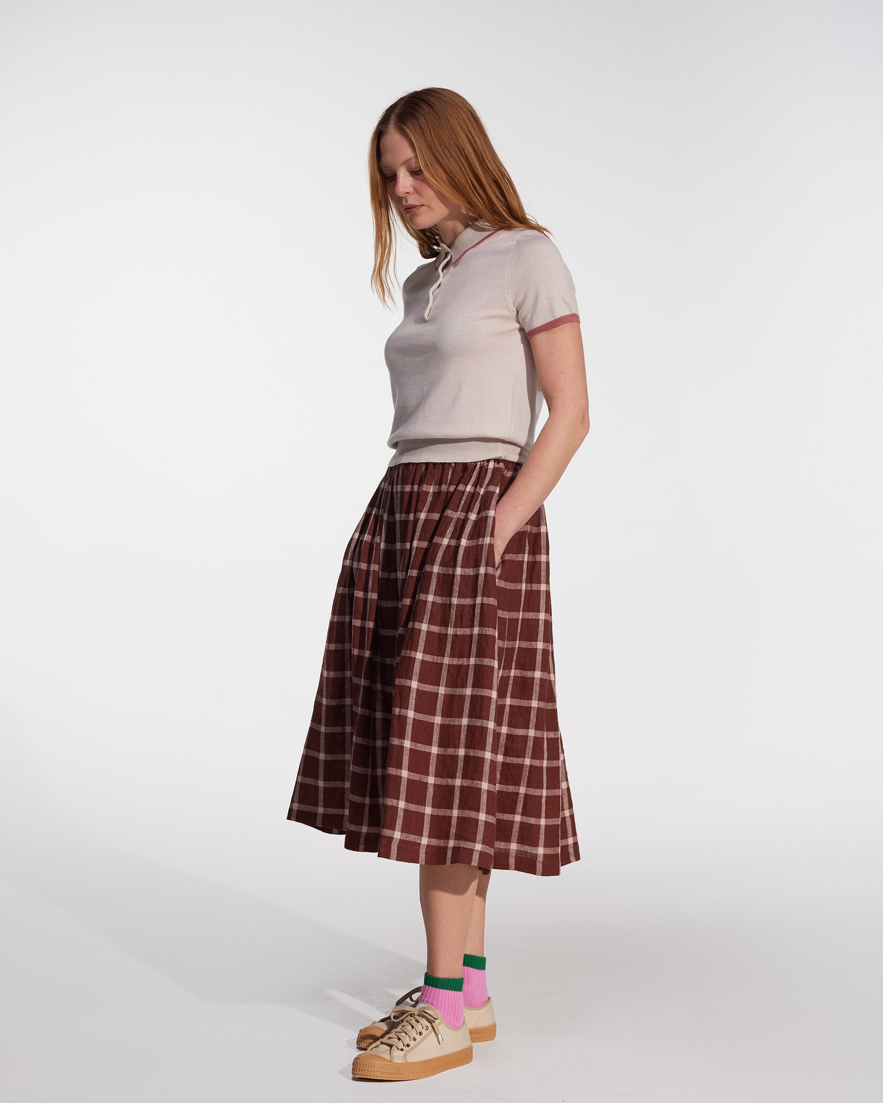 A woman stands with hands in pockets, wearing the Pyne and Smith Clothiers Linen Skirt No.11 in Neapolitan, a brown plaid European flax linen midi skirt with an elastic waist, paired with a beige top, beige sneakers, and pink socks against a plain background.
