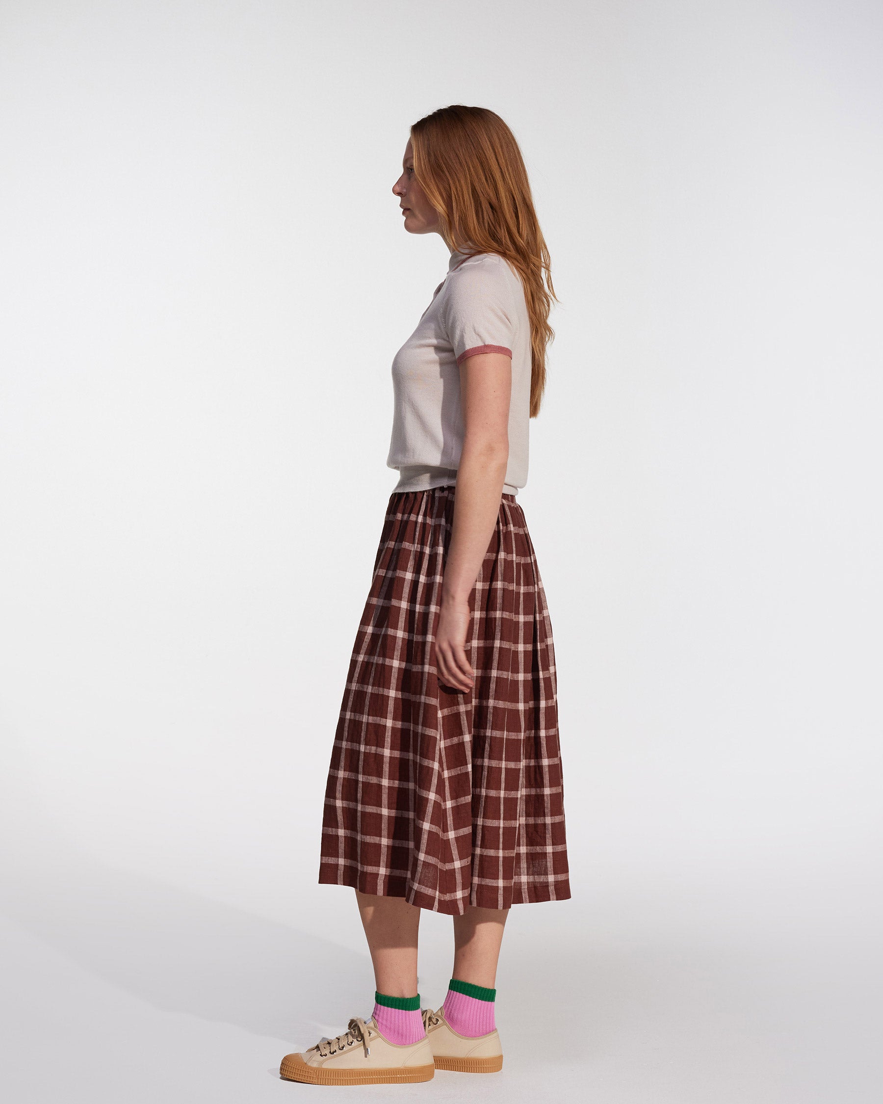 A woman with long red hair stands in profile against a plain background, wearing a light short-sleeve shirt and the Linen Skirt No.11 in Neapolitan by Pyne and Smith Clothiers. She pairs it with beige shoes and pink socks with green accents.