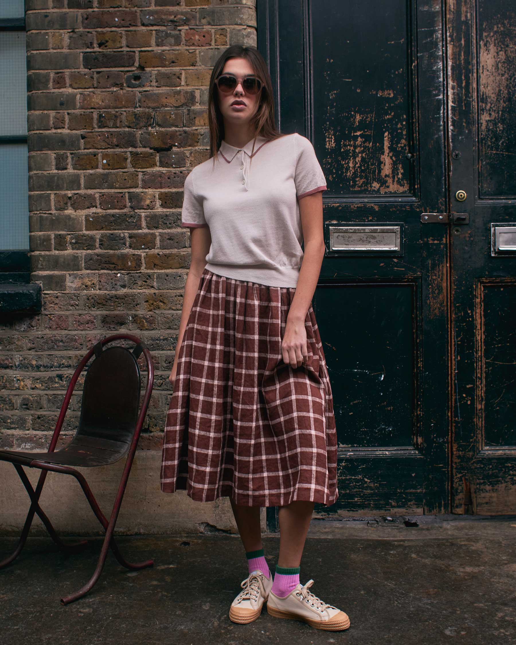 A woman stands outdoors by a brick wall and black door, wearing sunglasses, a white polo, pink socks, tan sneakers, and the Pyne and Smith Clothiers Linen Skirt No.11 in Neapolitan. A metal chair sits to her left.