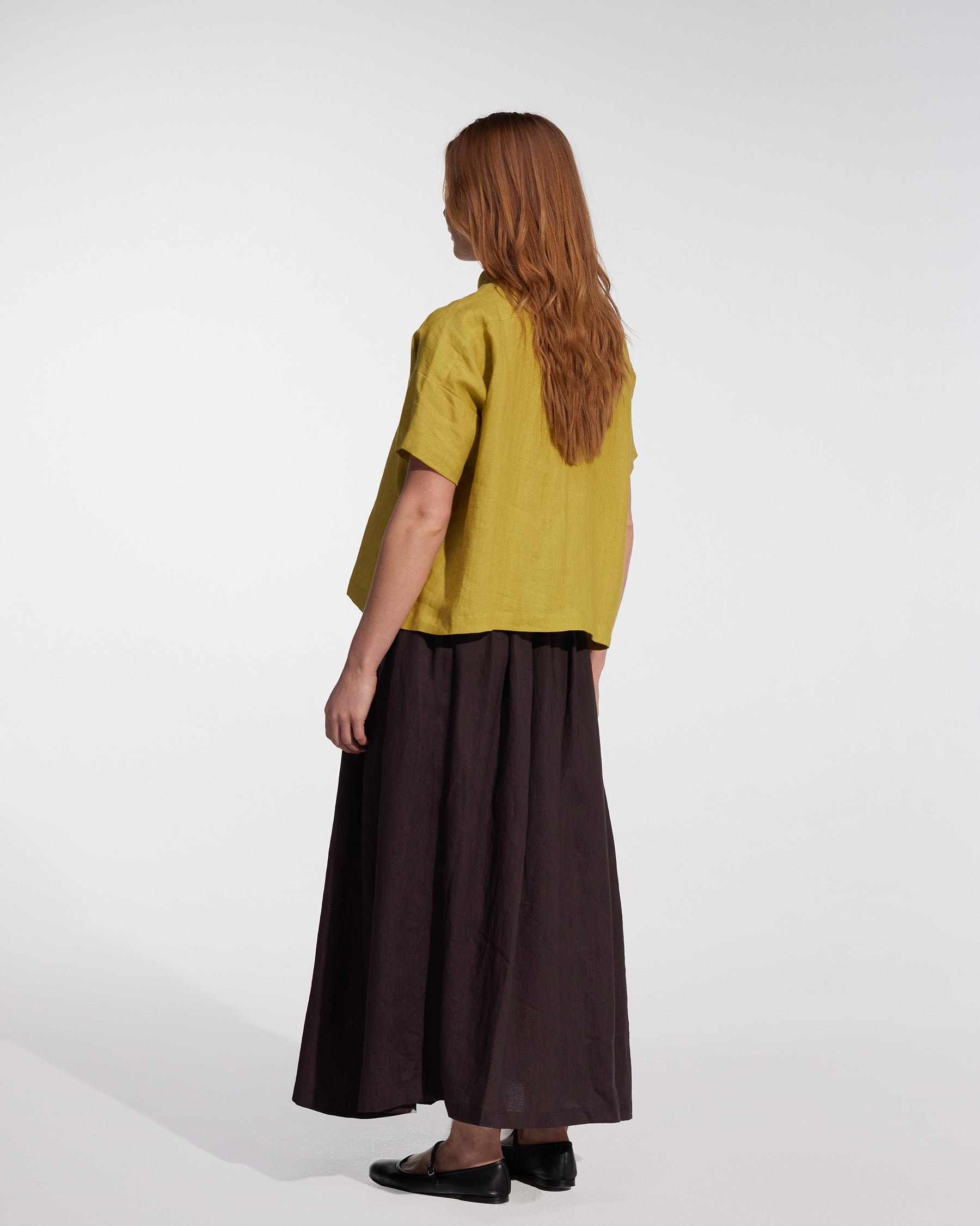 A person with long, light brown hair faces away, wearing a loose yellow short-sleeve top, black shoes, and the Linen Maxi Skirt No.12 in Chocolate Plum by Pyne and Smith Clothiers, standing against a plain white background.