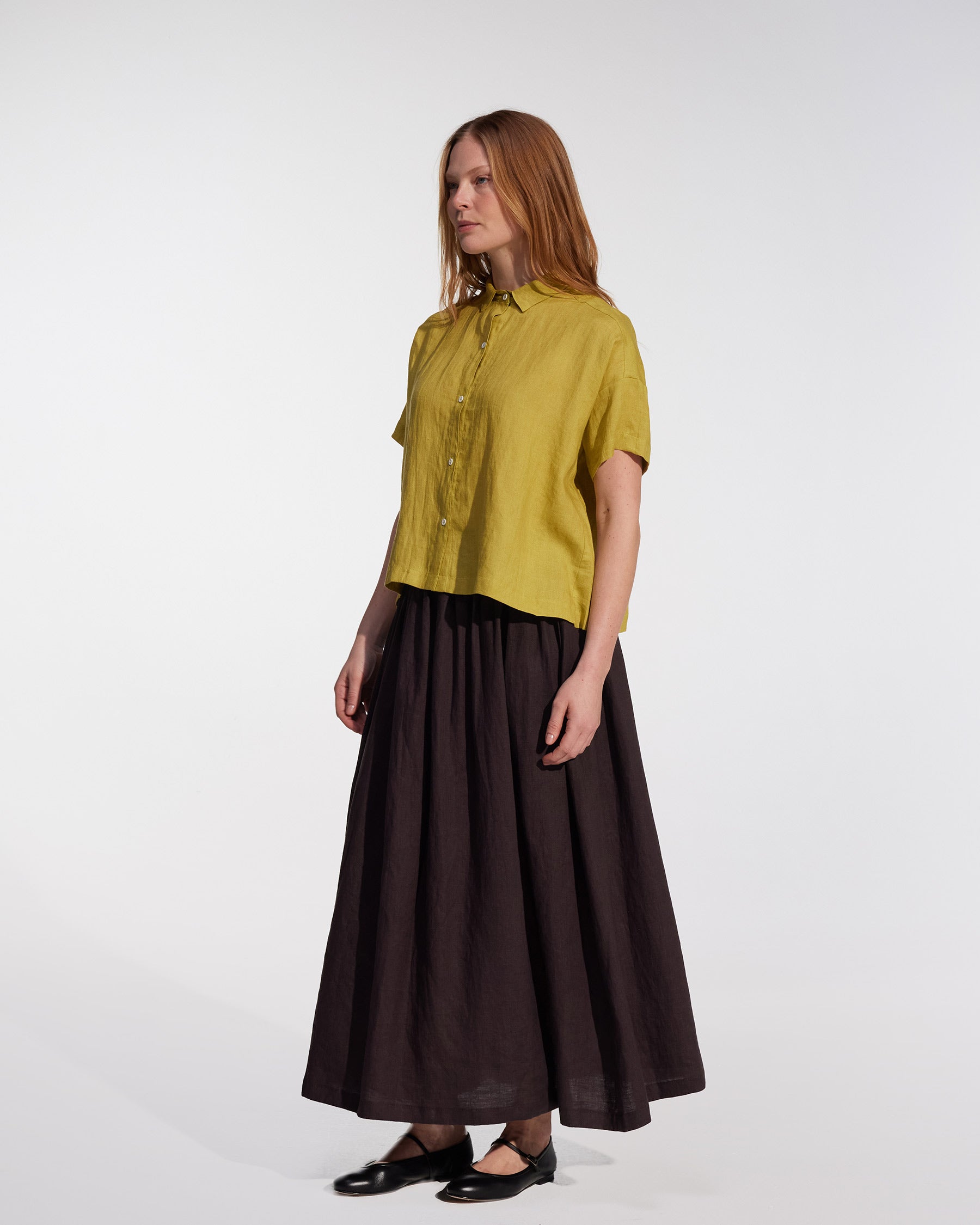 A woman with straight, light brown hair and a neutral expression wears a loose yellow blouse and the Pyne and Smith Clothiers Linen Maxi Skirt No.12 in Chocolate Plum, paired with black flats, against a plain white background.