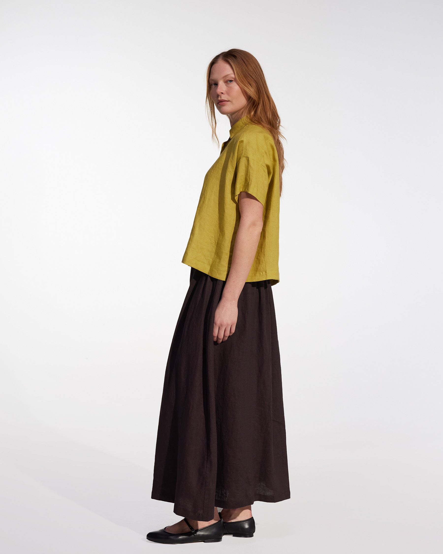 A woman with long red hair stands sideways against a white background, wearing a loose yellow short-sleeve top, black flats, and the Pyne and Smith Clothiers Linen Maxi Skirt No.12 in Chocolate Plum.