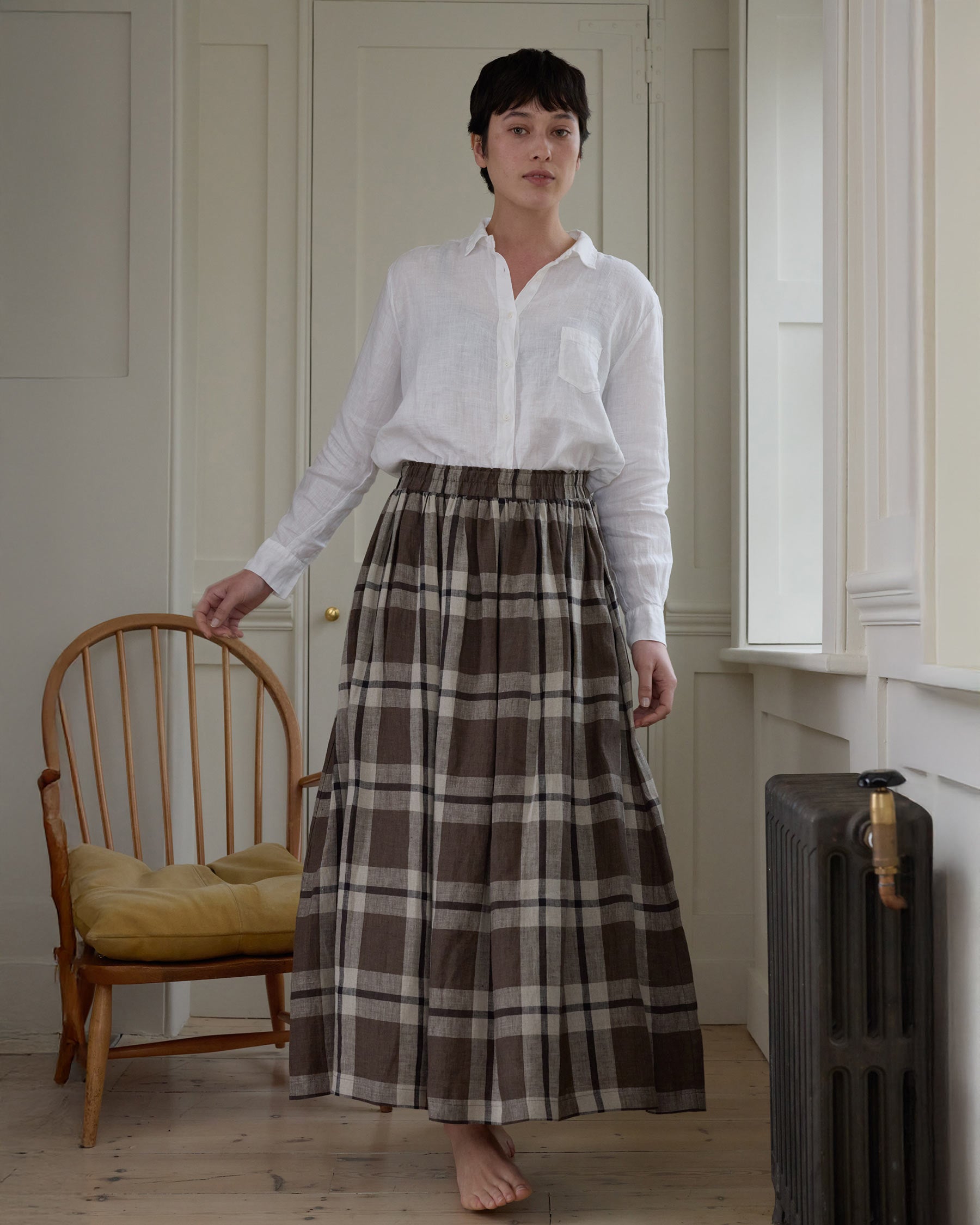 A person with short dark hair, barefoot and indoors, wears a white long-sleeve shirt and the Pyne and Smith Clothiers Linen Maxi Length Skirt No.12 in Fawkes Plaid, standing near a wooden chair with a yellow cushion and a radiator in soft light.