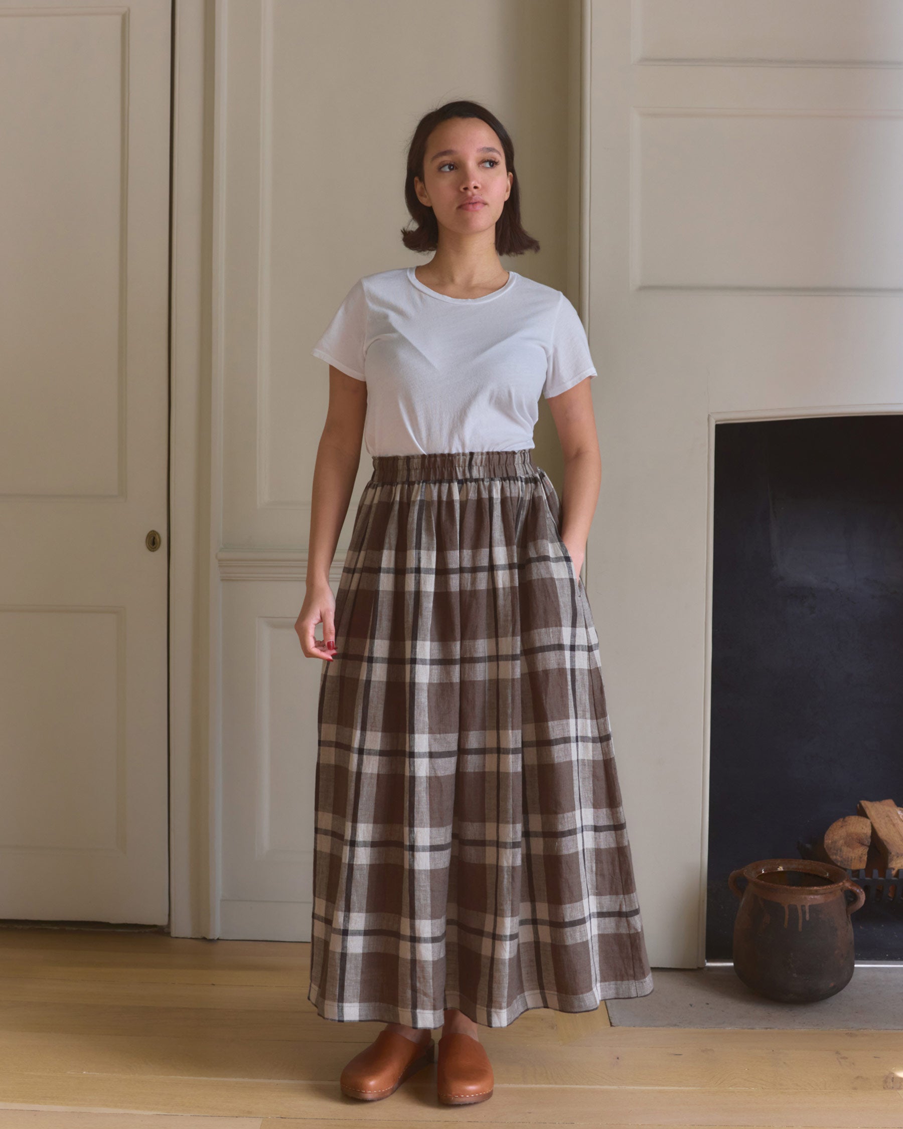 Indoors, a woman wears a white t-shirt, brown shoes, and the Linen Maxi Length Skirt No.12 in Fawkes Plaid by Pyne and Smith Clothiers. With one hand in her pocket, she gazes to the side; behind her are a fireplace and pottery.