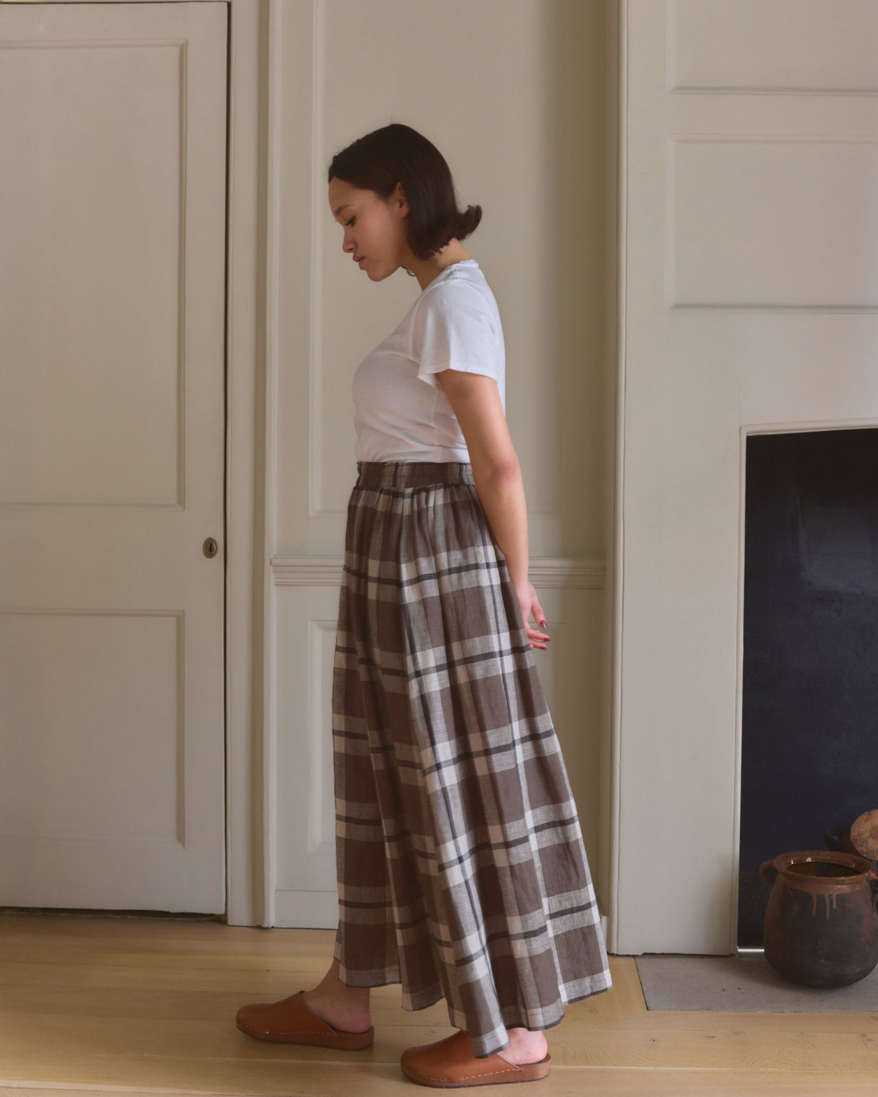 A woman stands indoors in profile, wearing a white t-shirt and the Pyne and Smith Clothiers Linen Maxi Length Skirt No.12 in Fawkes Plaid with brown slippers, hands behind her back as she looks down against a neutral background.