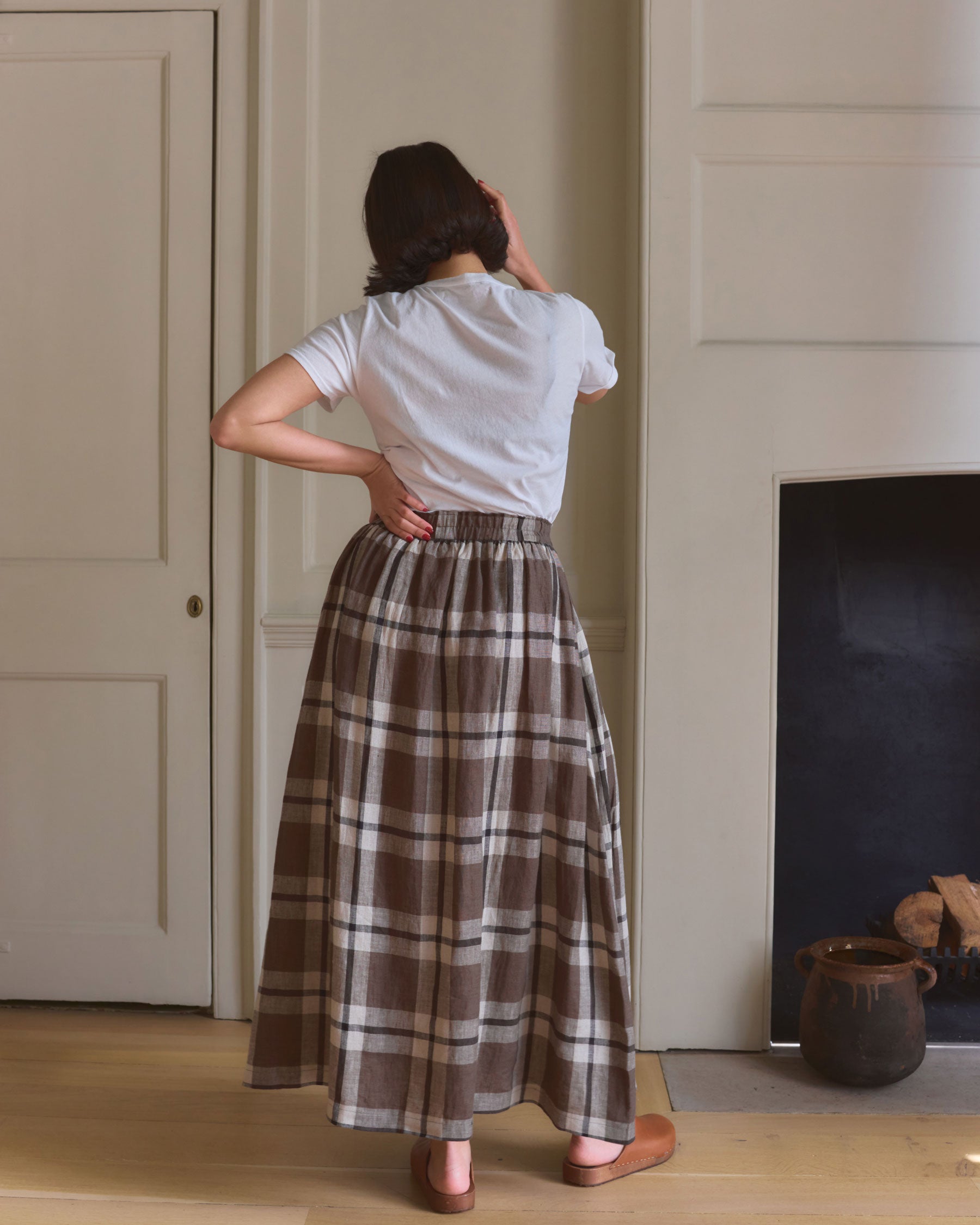 A woman stands indoors with her back to the camera, wearing a white short-sleeved top and the Pyne and Smith Clothiers Linen Maxi Length Skirt No.12 in Fawkes Plaid, near a fireplace and closed door, one hand raised to her head.