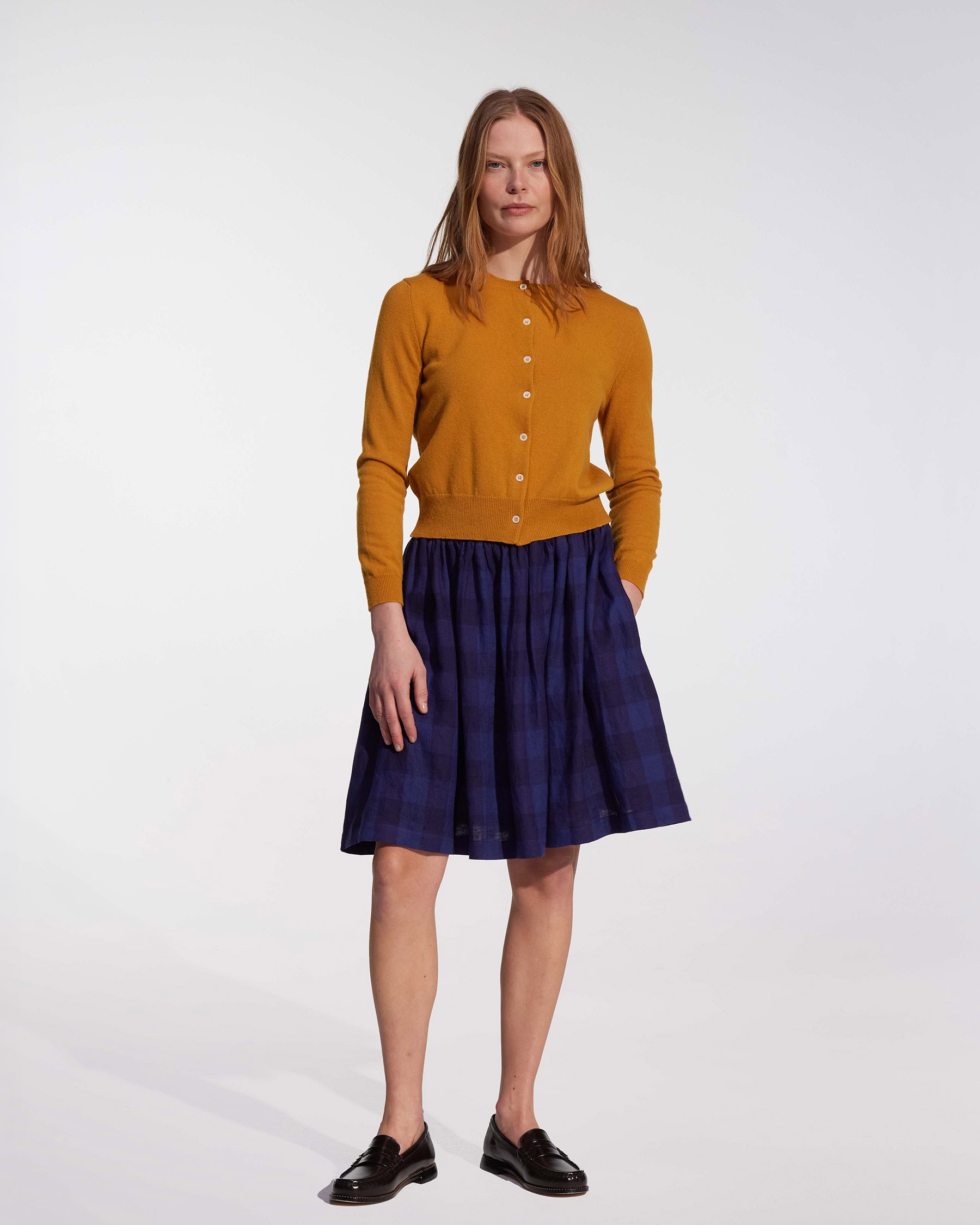 A woman with light brown hair and a neutral expression wears a mustard yellow cardigan, black loafers, and the Pyne and Smith Clothiers Linen Skirt No.14 in Midsommar Night Check against a plain background.
