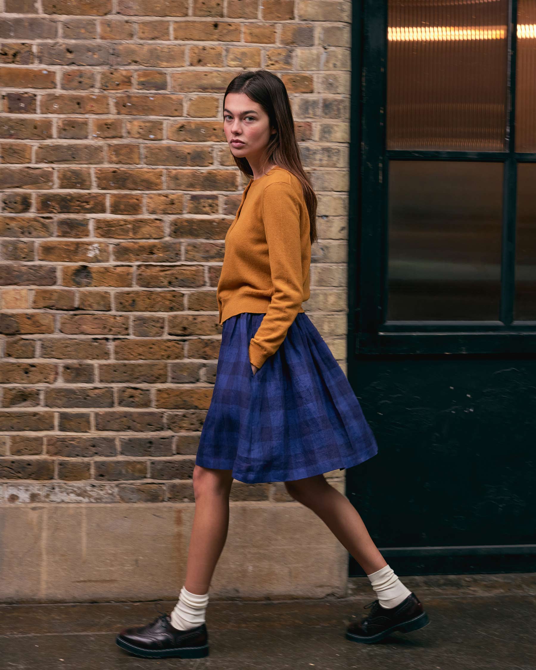 A woman in a mustard sweater, white socks, black shoes, and a Pyne and Smith Clothiers Linen Skirt No.14 in Midsommar Night Check walks past a brick wall and black door, looking towards the camera.