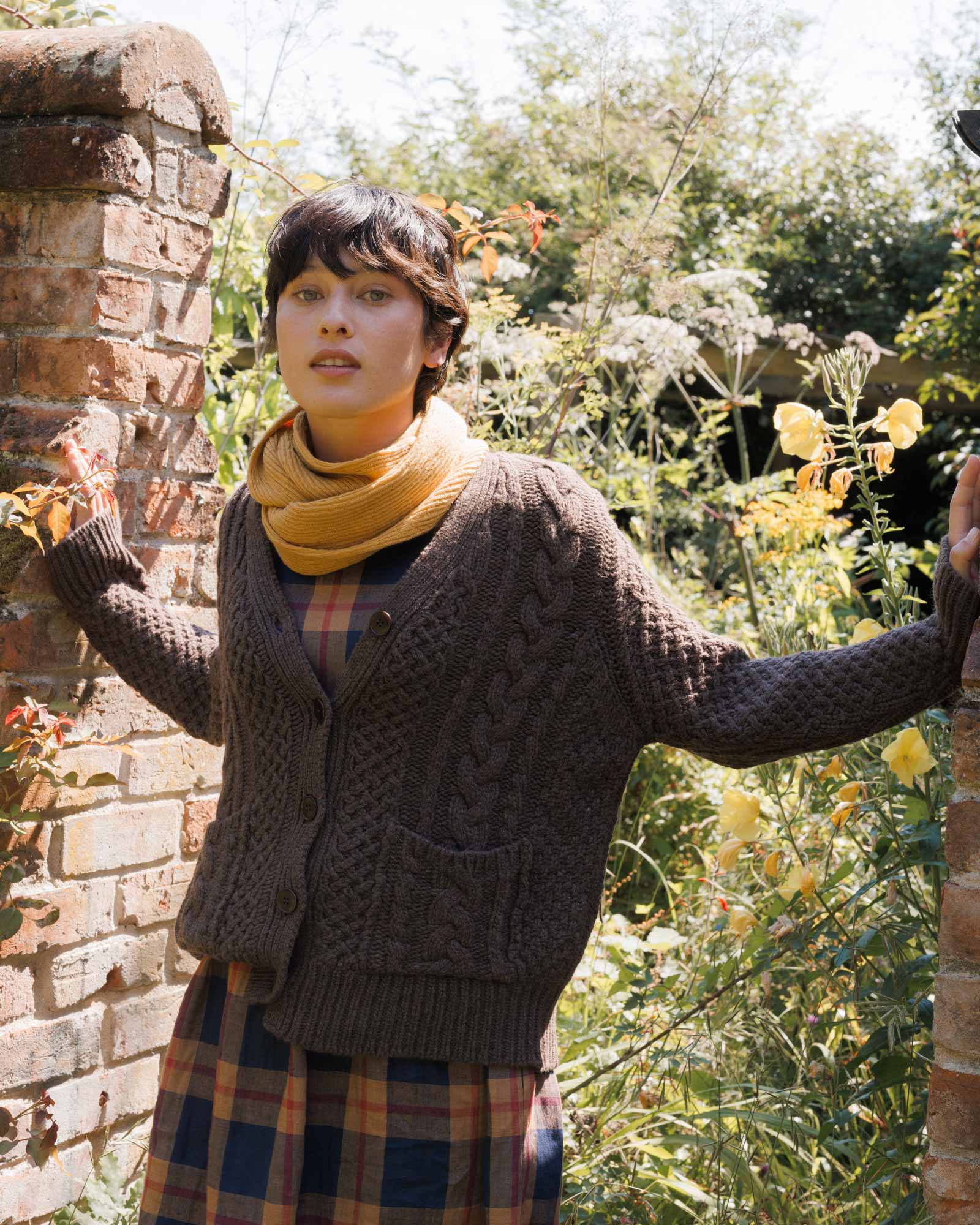 A person with short dark hair stands outdoors among greenery, wearing the Pyne and Smith Clothiers Merino Wool Cable Knit Cardigan with Pockets in Walnut over a plaid dress and mustard yellow scarf, between brick pillars in bright sunlight.