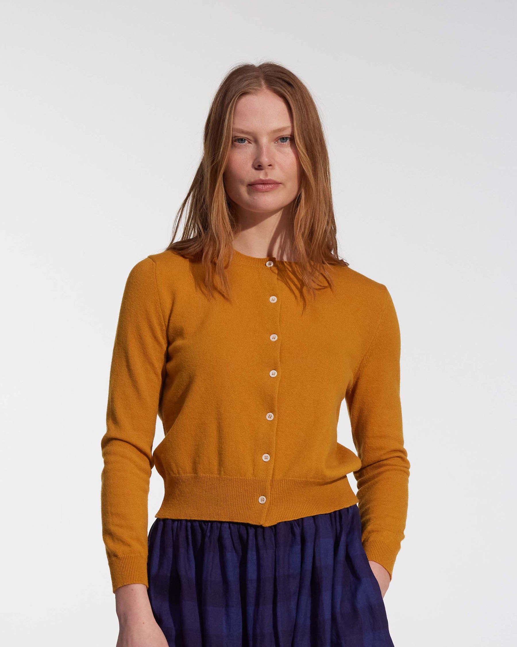 A woman with long reddish-brown hair wears the Pyne and Smith Clothiers Merino Cardigan in Coleman's Yellow and a dark blue plaid skirt, standing against a plain white background.