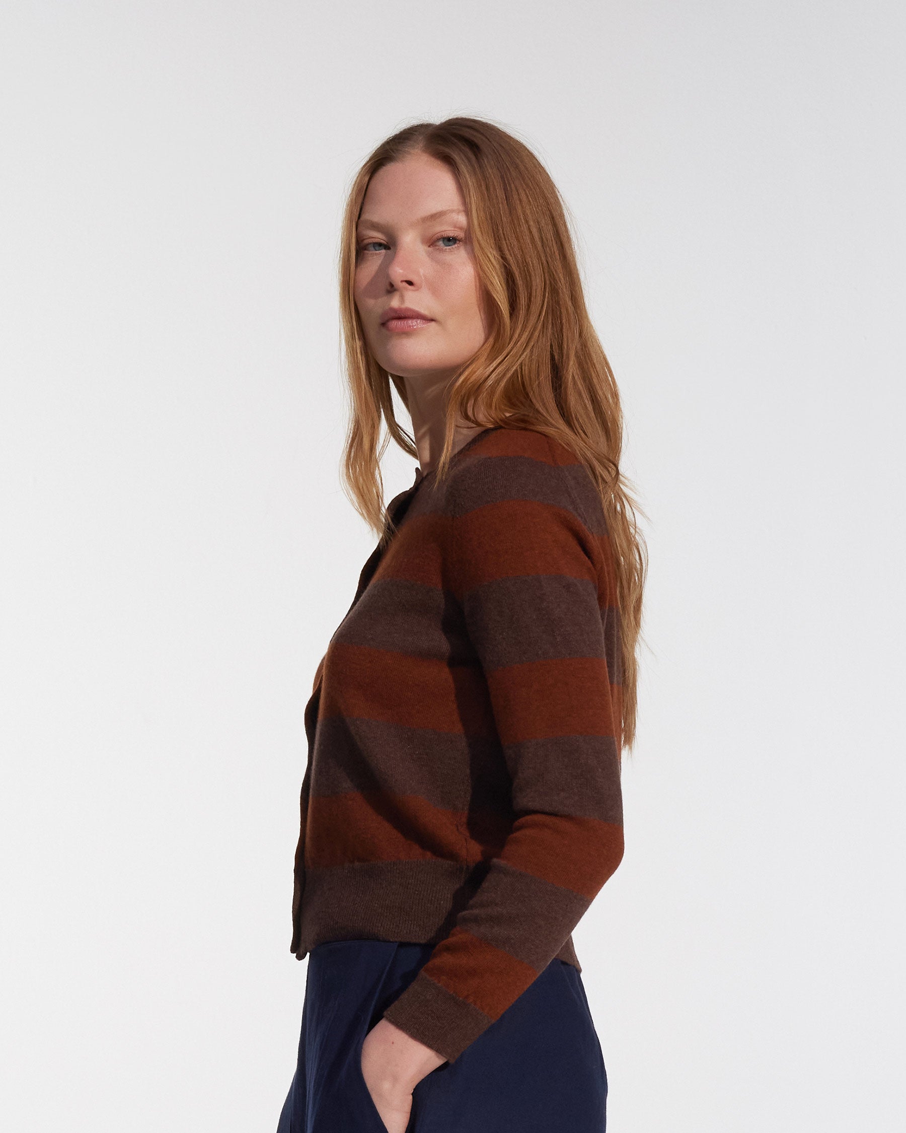 A woman with long, straight, light brown hair models the Pyne and Smith Clothiers Merino Cardigan in Foxtail, featuring brown and dark maroon stripes, with dark pants. She stands sideways and looks at the camera against a white background.