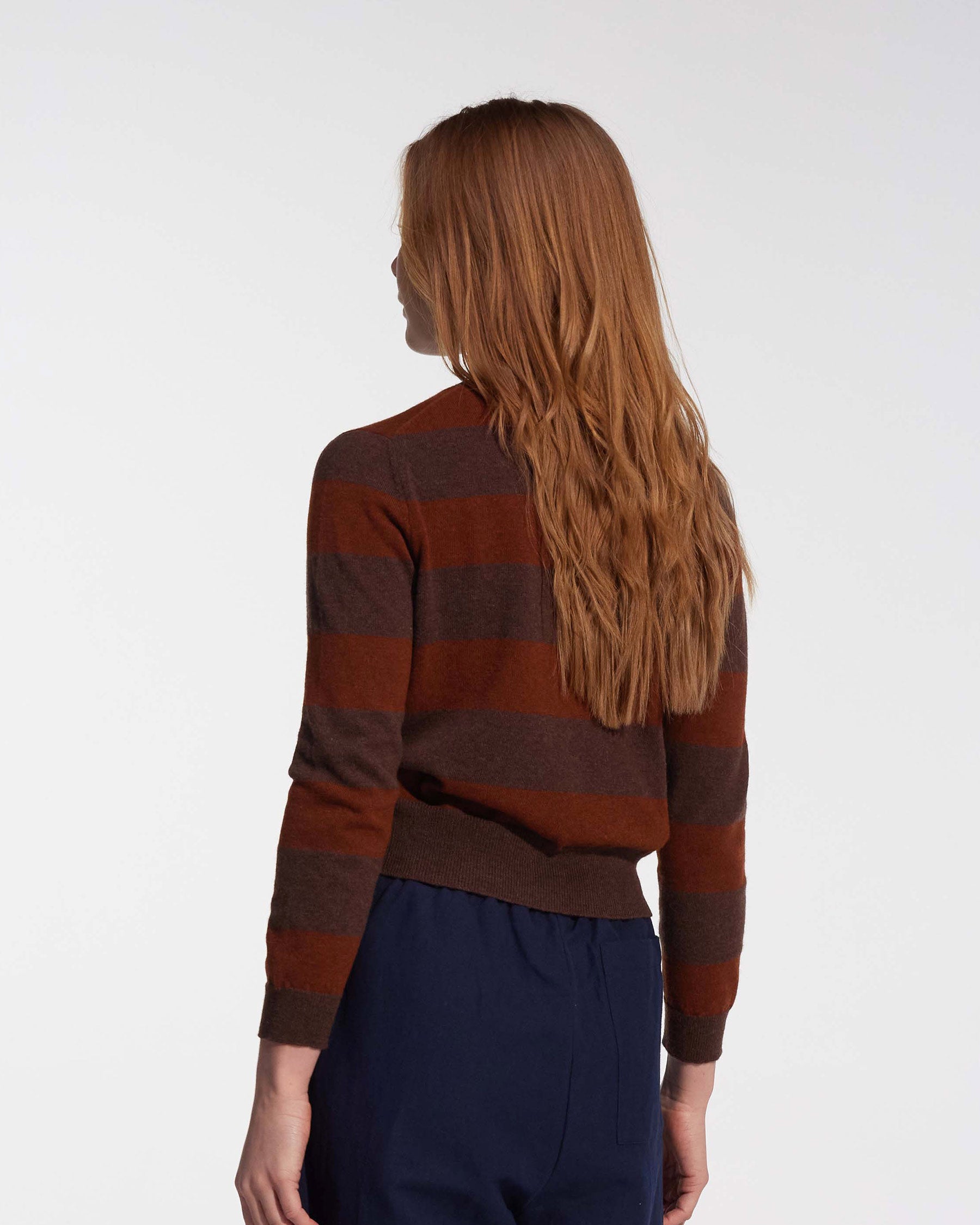 A woman with long, wavy auburn hair is seen from the back, wearing Pyne and Smith Clothiers’ Merino Cardigan in Foxtail with dark blue pants, standing against a plain white background.