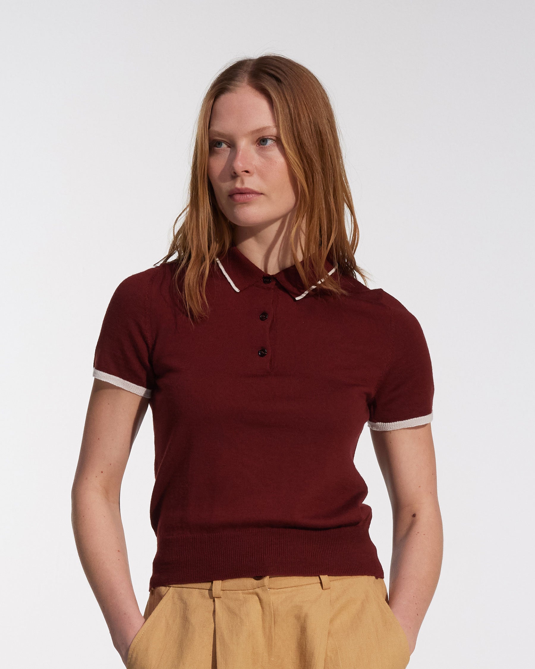 A woman with straight, shoulder-length reddish-brown hair models the Pyne and Smith Clothiers Merino Polo Shirt in Rosso, a slim-fitting Italian-made maroon top with light trim, paired with beige high-waisted pants against a plain background.