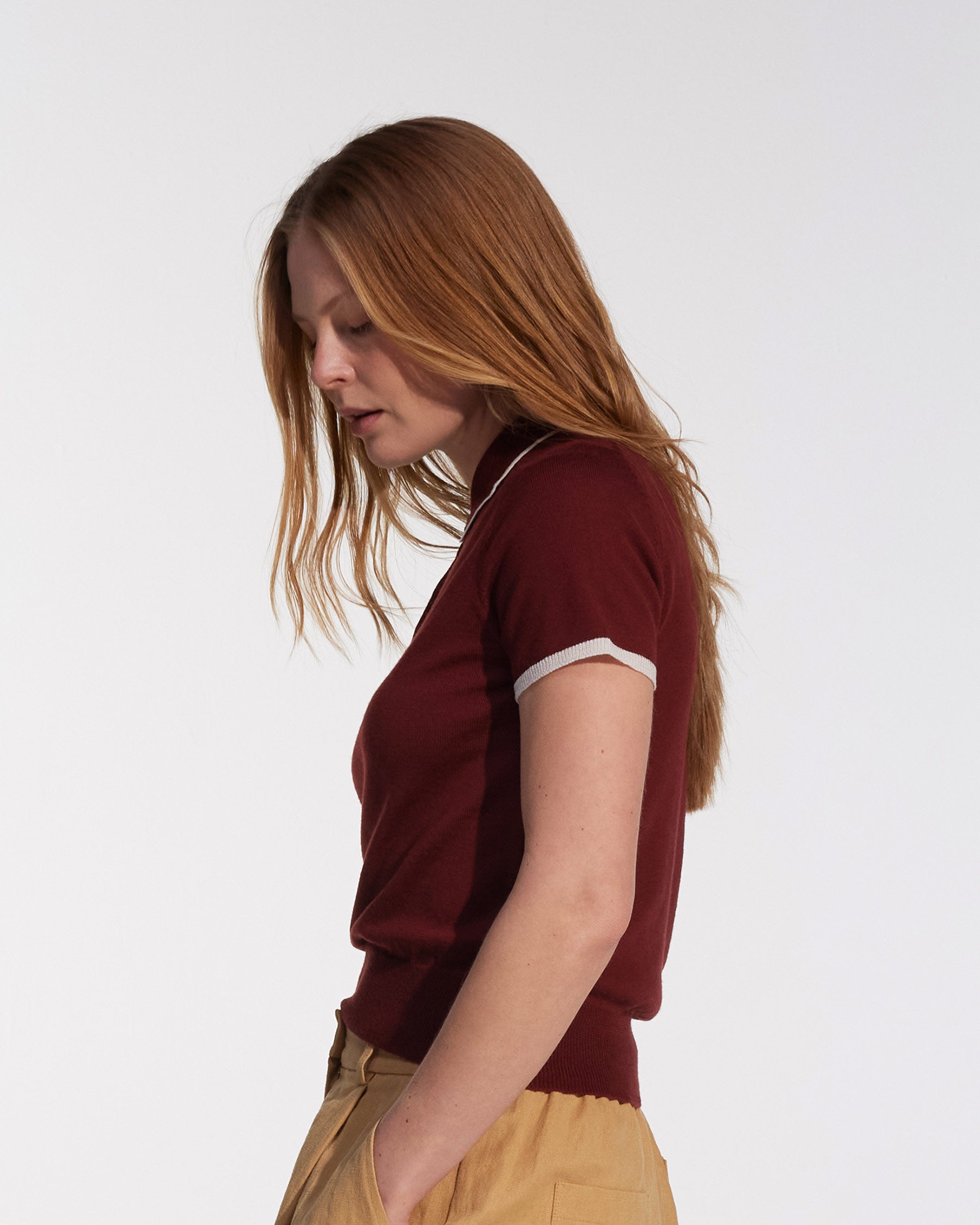 A person with long reddish-brown hair wears the Pyne and Smith Clothiers Merino Polo Shirt in Rosso and tan pants, standing in profile against a plain white background, looking down with a neutral expression.
