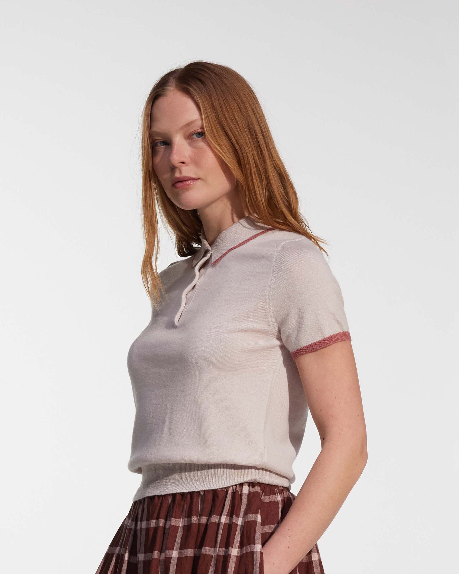 A woman with long red hair wears the Merino Polo Shirt in Shell Pink by Pyne and Smith Clothiers, a slim-fitting light knit with pink trim and a tie collar, paired with a brown checkered skirt, standing against a plain white background.